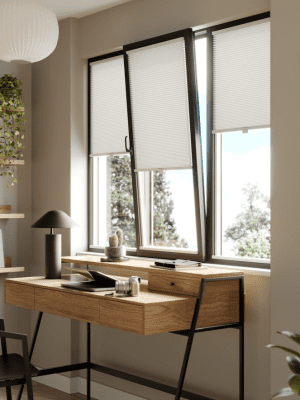 Clic Honeycomb This image shows white clic honeycomb blinds fitted onto a black frame tilt and turn window, above a wooden desk.